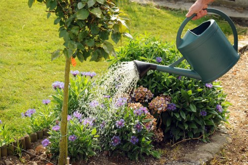 In a well-maintained backyard garden, a person waters a flower bed bordered by a small wooden edging, with a green watering can. The flower bed contains a variety of flowering plants, including purple and white blossoms, and is surrounded by lush green foliage. Behind the flower bed, a hedge or shrub provides natural privacy, while a grassy lawn extends in the background. The soil in the flower bed appears moist, indicating recent watering, and the garden is bathed in natural daylight, suggesting fair weather. The scene reflects typical outdoor garden maintenance activities, with attention to plant care and landscape organization, suitable for gardening services such as lawn mowing, planting, and watering.